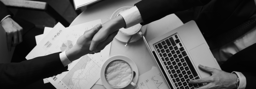 A black and white image of two professionals shaking hands over a business meeting table. The table is scattered with business reports, a laptop, and a coffee cup, symbolizing collaboration and modern B2B decision-making.